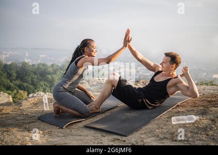 Joyeux couple divers en vêtements de sport donnant élevé cinq tout en ayant l'entraînement pour abs sur le sommet de la colline. Paysage urbain en arrière-plan. Modes de vie actifs. Banque D'Images