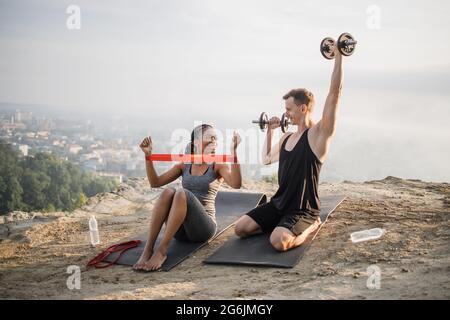 Femme africaine dans les vêtements de sport d'entraînement des bras en utilisant le caoutchouc de bande tandis que l'homme caucasien fort d'entraînement avec des haltères lourdes. Couple actif s'exerçant à l'extérieur. Banque D'Images