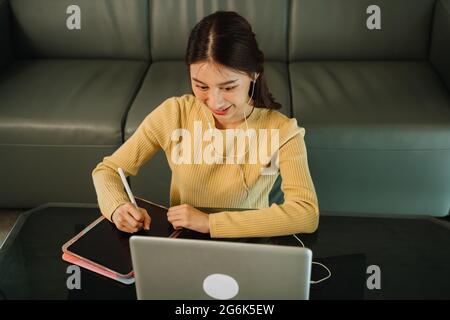 Vue en grand angle de la jeune femme d'affaires asiatique assistant à un appel vidéo avec écouteurs et écrivant des notes de réunion sur une tablette numérique tout en travaillant tard la nuit depuis chez elle Banque D'Images