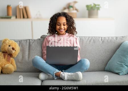 Communication et technologie. Jeune fille noire souriante assise sur un canapé et utilisant un ordinateur portable Banque D'Images