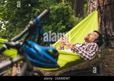 L'homme voyage à vélo, se détendre dans un hamac vert, surfer sur Internet sur un smartphone, écouter de la musique sur des écouteurs dans la forêt près du lac. Entrée cycliste Banque D'Images