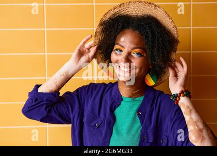 Portrait d'une femme souriante avec vitiligo dans un chapeau de paille vêtu de vêtements élégants près du mur orange du bâtiment Banque D'Images