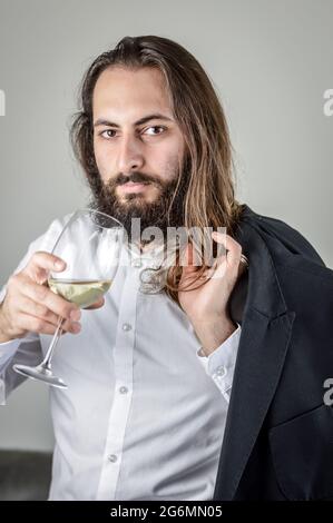 Portrait d'un jeune homme d'affaires du Moyen-Orient avec une barbe et des cheveux longs tenant un verre de vin blanc et sa veste sur son épaule Banque D'Images
