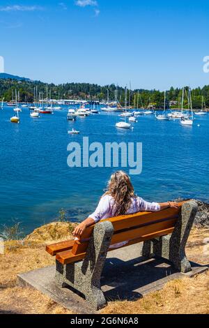 Femme aux cheveux blonds touffés assise sur un banc qui regarde Shoal Harbour, à Sidney, en Colombie-Britannique, au Canada Banque D'Images