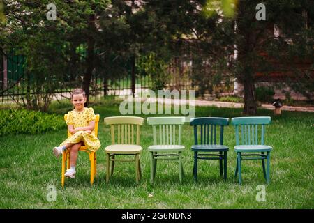 Une fille rieuse et gaie dans une robe jaune est assise sur des chaises viennois peintes multicolores attendant un ami pour une fête d'enfants et un anniversaire Banque D'Images