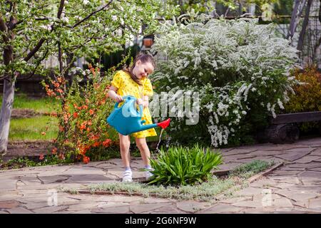 une fille heureuse, riante dans une robe jaune est arroser des fleurs d'un arrosoir bleu, un jeune jardinier est jardinage dans l'arrière-cour avec la floraison Banque D'Images