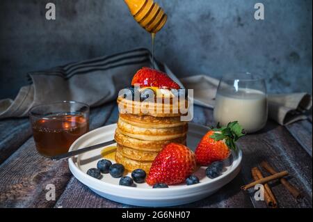 Une pile de crêpes moelleuses avec du miel, des myrtilles fraîches, des fraises et un verre de lait sur une table en bois, délicieux dessert pour le petit déjeuner Banque D'Images