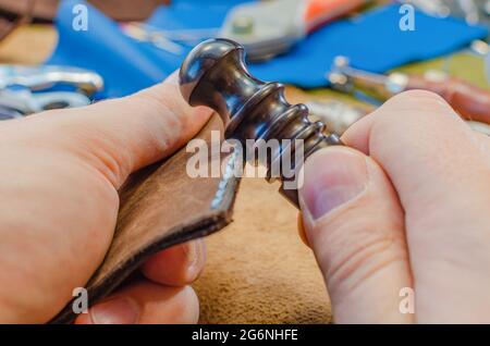 L'artisan grince le bord du produit en cuir. Les outils et les accessoires sont répartis sur toute la table. Fabrication de produits en cuir. Banque D'Images