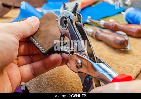 Le maître tient un morceau de cuir avec un perforateur. Les outils et les accessoires sont répartis sur toute la table. Banque D'Images