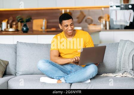 Beau et positif jeune latino-homme dans des vêtements décontractés, assis sur le canapé à la maison dans le salon, à l'aide d'un ordinateur portable, la navigation sur Internet, le chat sur les réseaux sociaux, regarder l'écran, sourires Banque D'Images