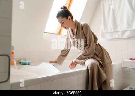 Jeune femme blanche souriante en peignoir se pliant au-dessus de la baignoire remplie de bulles, en testant la température de l'eau Banque D'Images