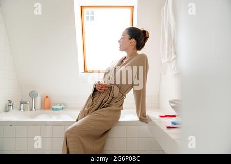 Jeune femme blanche souriante en peignoir se pliant au-dessus de la baignoire remplie de bulles Banque D'Images