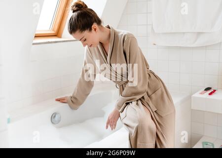 Jeune femme blanche souriante en peignoir se pliant au-dessus de la baignoire remplie de bulles, en testant la température de l'eau Banque D'Images
