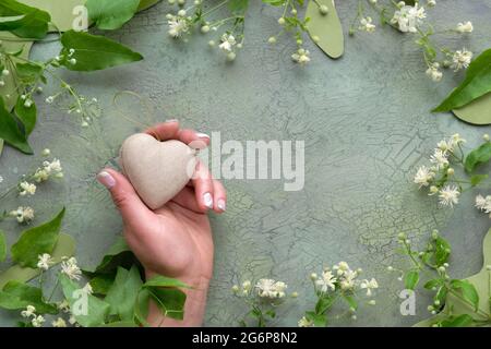 Main tenant le coeur de papier recyclé d'artisanat. Plat avec fleurs blanches sauvages et feuilles vertes. Cadre naturel sur vert peint texturé vieilli Banque D'Images