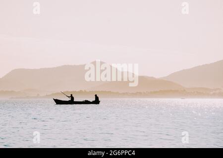 Silhouette de pêcheurs sur un petit bateau à la mer contre des montagnes lumineuses en arrière-plan Banque D'Images