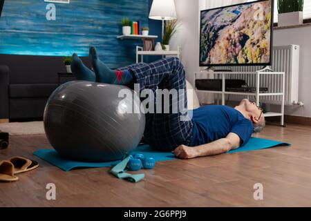 Retraite homme senior faisant bien-être réchauffant les jambes à l'aide de ballon suisse assis sur le tapis de yoga dans le salon. Retraité retraité exerçant abdominaux exercices de travail à la santé musculaire de corps Banque D'Images