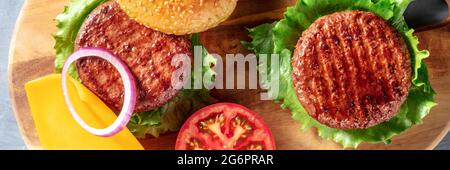 Panorama Burger avec deux hamburgers dans le processus de fabrication dans la cuisine, avec du fromage et des légumes, tiré du dessus sur une planche en bois Banque D'Images