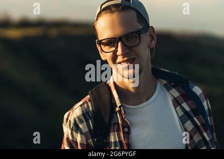 Portrait d'un jeune homme beau, portant des lunettes et une casquette, jeune homme souriant, debout dans la rue dans la nature, sur le fond d'un beauffu Banque D'Images