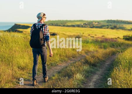 Souriant jeune homme élégant, portant des lunettes et une casquette, voyageant avec un sac à dos en été, randonneurs et observant le paysage pittoresque, Trave Banque D'Images