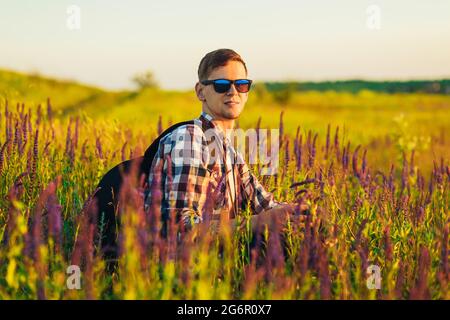 Jeune homme en lunettes de soleil assis dans un champ avec des fleurs sauvages, homme à la campagne pendant le coucher du soleil, appréciant la nature, concept de voyage, nature Banque D'Images