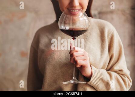 Crop Happy female en chandail sentant le vin rouge dans un grand verre sur fond gris Banque D'Images