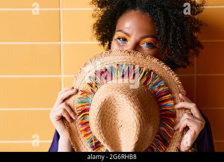 Femme afro-américaine avec chapeau de paille coloré devant le visage ayant un maquillage brillant et élégant debout près du mur orange et regardant l'appareil photo. Bonjour Banque D'Images