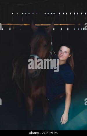 Horsewoman posant avec son cheval brun de phoque dans l'écurie. Fille tenant l'étalon avec affection. Liaison entre êtres humains et chevaux concept. Banque D'Images