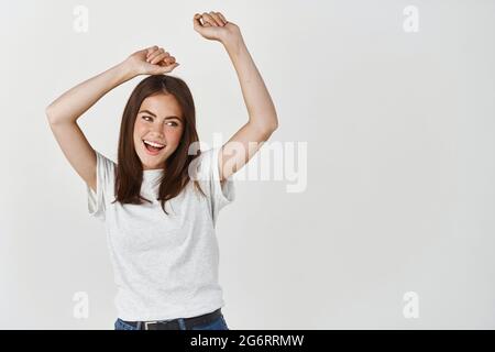 Belle femme brune s'amusant, dansant à l'intérieur avec les mains levées et souriant, debout heureux sur fond blanc Banque D'Images