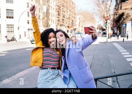 De belles jeunes filles avec des cheveux bouclés souriant et prenant selfie avec téléphone mobile dans la rue. Banque D'Images