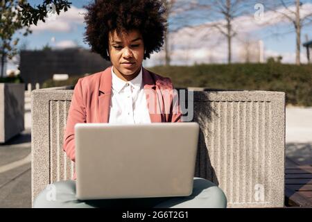 Femme d'affaires avec cheveux afro portant des vêtements formels assis à l'extérieur et utilisant son ordinateur. Banque D'Images
