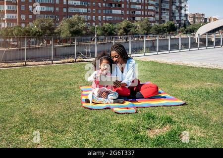 Bonne petite fille avec des cheveux afro assis dans le parc avec sa mère et en utilisant une tablette pendant la journée ensoleillée. Banque D'Images