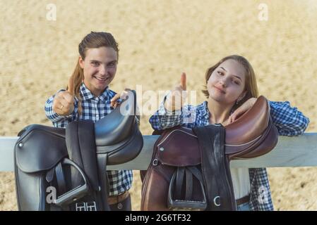 Deux jeunes filles debout dans l'arène de sable pour l'équitation. Les filles de bonne humeur et de faire des pouces signe vers le haut à l'appareil photo. Selles en cuir suspendues sur la clôture en bois au premier plan. Banque D'Images