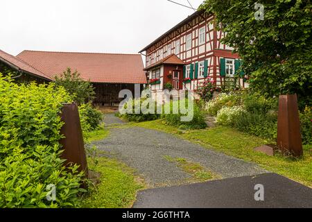 Une ferme en colombage bien entretenue avec un toit de tuiles rouges, des roses en fleurs et une grange ouverte se dresse près d'une entrée de ferme à Görsdorf, hameau de la ville de Schalkau en Thuringe, Allemagne. Pendant la division allemande, Görsdorf se trouvait dans la zone frontalière interdite de 500 mètres de la RDA et a été entourée en 1981 par un mur de béton d'environ 3,5 mètres utilisé comme écran visuel. Banque D'Images