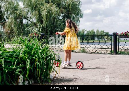 vue arrière d'une fille dans une robe jaune à cheval un scooter sur un chemin avec des lits de fleurs vertes, une fille apprenant à monter un scooter le long d'une rivière en été Banque D'Images