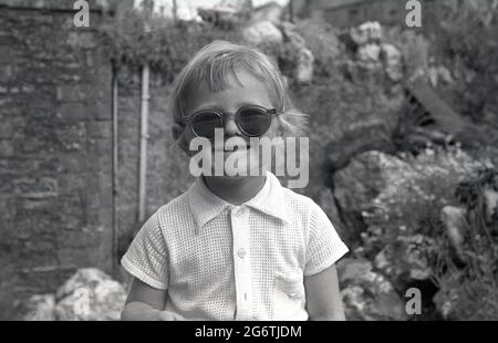 Années 1960, historique, à l'extérieur dans un jardin de cour, une petite fille portant une groovy paire de lunettes de soleil, Angleterre, Royaume-Uni. Banque D'Images