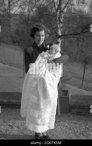 Années 1950, historique, une nouvelle mère debout à l'extérieur tenant son bébé enveloppé dans un long coton ou de dentelle à motif châle, à l'occasion spéciale du baptême du bébé, Angleterre, Royaume-Uni. Les châles de baptême sont des vêtements de fête utilisés lors de la cérémonie spéciale lorsque l'eau « sainte » est versée sur la tête du bébé. Banque D'Images