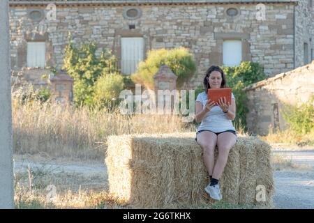 Scène rurale avec brunette d'âge moyen latina femme consultant une tablette avec couverture orange assis sur une balle de paille avec maison en pierre dans l'arrière-plan. Banque D'Images