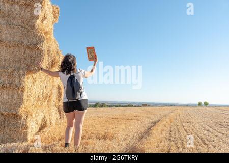 Brunette Latina d'âge moyen, femme debout consultant une tablette penchée sur une pile de balles de paille au milieu d'un champ de céréales récolté. Debout Banque D'Images