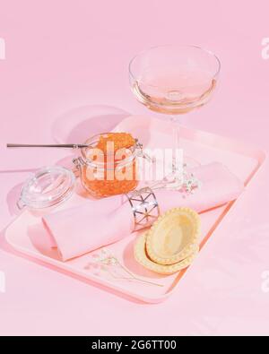 Vertical, élégant, rose, composition alimentaire avec un pot de caviar rouge de saumon, tartelettes et un verre de vin rose sur un plateau sur fond rose Banque D'Images