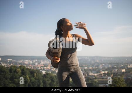 Belle femme de grande taille avec la peau noire boire de l'eau douce à partir d'une bouteille après l'entraînement à l'extérieur. Jeune femme en vêtements de sport rafraîchissants après l'entraînement. Banque D'Images