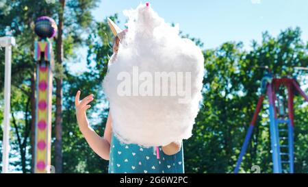 Un enfant mange une énorme quantité de bonbons en coton dans un parc d'attractions en été. Concept d'enfance heureux et insouciant. Bonbons de coton dans les mains d'un enfant proche- Banque D'Images