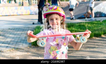 Une fille dans un casque tient une planche à roulettes dans ses mains. Le skateboard pour les enfants et les adolescents dans le parc des sports extrêmes pendant les vacances d'été. Actif Banque D'Images