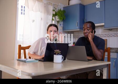 Un couple travaille à la maison sur l'ordinateur, la femme entend ce que son mari dit pendant une conversation au téléphone. Couple interracial Banque D'Images
