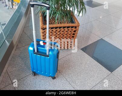 Une valise bleue à roulettes, située sur un sol en marbre dans un terminal moderne d'aéroport ou de gare, sur le fond d'un grand pot en osier Banque D'Images