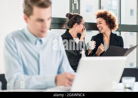 Deux jeunes femmes de rire tout en parlant dans le bureau derrière leur collègue masculin Banque D'Images