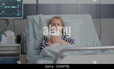 Portrait d'un petit enfant malade solitaire qui se repose dans un lit tenant un ours en peluche dans les mains, regardant la caméra pendant l'examen de récupération à l'hôpital. Enfant hospitalisé en attente de soins de santé Banque D'Images