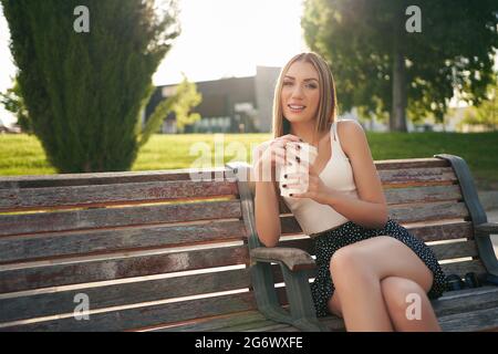 Belle femme assise sur un banc de parc dans des vêtements d'été ayant un café regardant l'appareil photo. Photo de haute qualité Banque D'Images