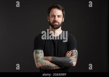 Barbu brunette homme avec tatouage posant et regardant la caméra isolée sur fond noir Banque D'Images