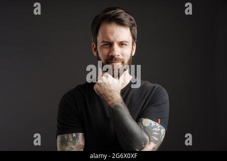 Barbu brunette homme avec tatouage posant et regardant la caméra isolée sur fond noir Banque D'Images