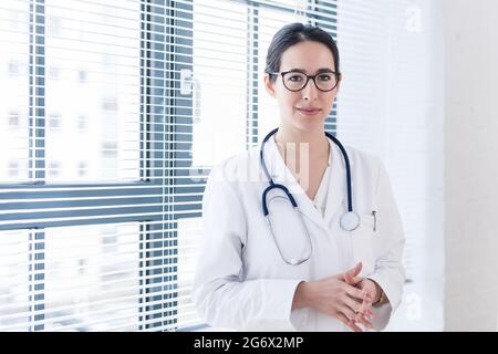 Portrait d'une jeune infirmière ou médecin portant des lunettes et une blouse médicale blanche pendant la recherche de caméra avec confiance et sérénité à l'intérieur Banque D'Images